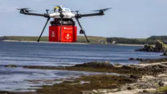 Drone branco da Skyports carrega uma caixa vermelha com o logotipo do correio britânico, sobre uma praia, com o mar ao fundo