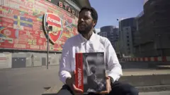 Dr Clive Chijioke Nwonka tenant un exemplaire du livre Black Arsenal devant l'Emirates Stadium