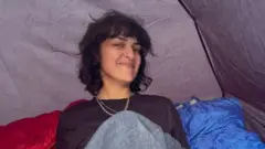 Henrietta, uma mulher de cabelos castanhos curtos e franja, sorri. Ela está sentada em uma barraca, vestindo uma blusa preta de mangas compridas e dois colares.
