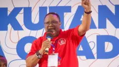 Umunyapolitike wo muri Tanzania Tundu Lissu arimo kuvugira mu ndangururamajwi. Yambaye umupira w'amaboko magufi w'umutuku, yazamuye igipfunsi. 

