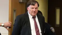 Rex Heuermann mira a la cámara con gesto desafiante. Viste un traje oscuro, camisa blanca y corbata granate. Se ve una mano de una persona que sujeta una puerta detrás de él
