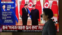 Una mujer pasa junto a una pantalla de televisión que muestra un noticiero con imágenes de archivo de la reunión de 2019 entre el presidente chino Xi Jinping y el líder norcoreano Kim Jong Un, en una estación de tren de Seúl el 8 de junio de 2026. 