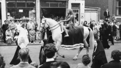 Una mujer desnuda y con el cabello muy largo pasea en un caballo en un desfile por la calle de Reino Unido. Hay personas mirando y varias monjas acompañan el desfile. La foto es en blanco y negro.