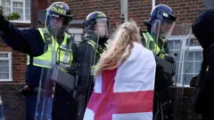 A woman with an England flag around draped around her is surrounded by policemen holding shields 