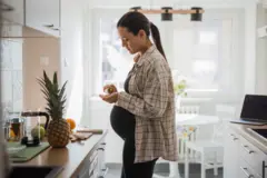 Una mujer embarazada, parada frente al estante de una cocina saca una pastilla de un frasco
