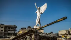 صورة تظهر تمثالاً أمامه دبابة مدمرة وسط ميدان في مدينة كوباني أو عين العرب شمالي سوريا.