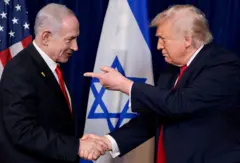 Benjamin Netanyahu e Donald Trump apertam as mãos enquanto estão em frente às bandeiras dos EUA e de Israel. Ambos os homens estão vestindo ternos azuis escuros com gravatas vermelhas, enquanto Trump está apontando para Netanyahu.
