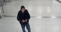 Johannes Natland usa moletom com capuz, jaqueta escura e calça jeans. Ele está em um aeroporto segurando um celular