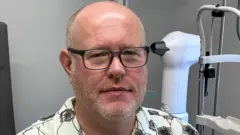 Un homme portant des lunettes et une chemise blanche à fleurs noires se trouve dans une salle d'un cabinet d'opticiens.