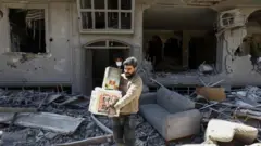 Dois homens em meio aos escombros de uma casa severamente destruída, com paredes perfuradas, janelas quebradas e móveis revirados. Em primeiro plano, um homem carrega uma caixa, enquanto outro, parcialmente ao fundo e usando máscara, o acompanha. Ao redor deles, há destroços espalhados, incluindo um sofá.