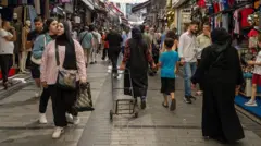 Çarşıda alışveriş yapan insanlar mağazaların arasından geçiyor. 