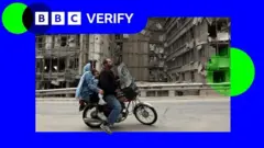 یک مرد و زن و کودکی خردسال سوار بر موتور از مقابل ساختمان‌های آوار عبور می‌کنند، لوگوی بی‌بی‌سی وریفای در گوشه تصویر دیده می‌شود