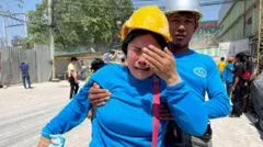 Una mujer llora mientras un hombre la sostiene, ambos visten camisetas azules y portan cascos protectores.