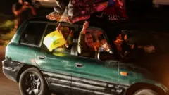 緑色の乗用車に乗る家族が、カメラに向かって笑顔とこぶしを見せて喜びを表している。車の後部座席や屋根の上には荷物が積まれている