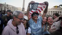 Grupo de pessoas comemorando na Praça São Pedro — um homem está segurando um terço e a bandeira dos EUA