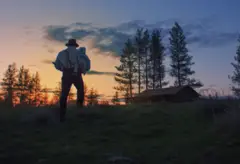 A imagem mostra a silhueta de um homem caminhando por um campo ao entardecer, carregando nos braços uma criança envolta em um pano claro. Ele usa chapéu, camisa de mangas compridas com suspensórios e calças escuras. Ao fundo, o céu está tingido por tons de laranja e azul do pôr do sol, com nuvens espalhadas e a silhueta de árvores altas. À direita, há uma casa de madeira simples, parcialmente envolta pela penumbra, reforçando a atmosfera rural e contemplativa da cena.