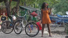 Una mujer sostiene su teléfono móvil en medio de bicicletas en una zona costera de Puerto Viejo. En el lugar hay personas locales y turistas que se congregan alrededor de un restaurante tradicional donde una banda toca música de calipso en vivo.