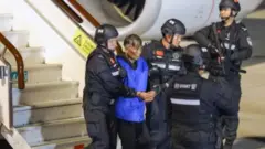 Một người đàn ông lớn tuổi mặc áo vest màu xanh đang bị cảnh sát dẫn giải ra khỏi máy bay