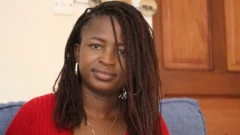 Noella sits inside facing the camera with a small smile. She wears silver hoop earrings, a necklace and a red top. 