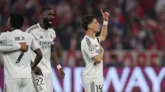 Arda güler ayakta, gözleri kapalı, sol elini kalbine koymuş, sağ elinin işaret parmağı gökyüzünü gösteriyor. 