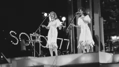 Duas mulheres com vestidos claros cantam no palco com microfones. Ao fundo, lâmpadas formam o logotipo da edição de 1977 do festival Intervision, realizado na cidade polonesa de Sopot. A imagem é em preto e branco.