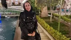 زهرا طبري په یوه پاک کې په تورو کلیو کې ولاړه ده چې تر څنګ یې اوبه هم بهېږي او شاوخوا یې د پارک بوټي او چمن هم برېښي.