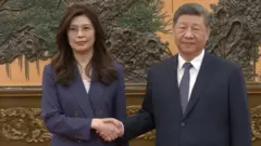 Cheng Li-wun, chairperson of the Kuomintang (KMT), Taiwan's largest opposition party, shakes hands with Chinese President Xi Jinping at the Great Hall of the People in Beijing, China April 10, 2026, in this screengrab from a video provided by CTI. 木彫りの模様がついた壁を背に、ともに紺色のスーツを着た鄭氏と習氏が握手している。