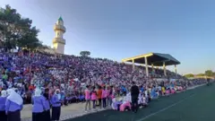 ዝተፈላለየ ሕብሪ ዝተኸድነ ህዝቢ ኣብ ደረጃታት ሜዳ ኩዕሶ እግሪ ኮፍ ኢሎም፡ ብድሕሪት ካብ ሜዳ ወጻኢ ሓደ ናብቲ ብሩህ ሰማይ ዝወርወር መስጊድ ይርአ