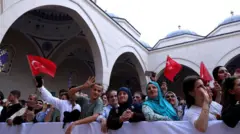 Başörtülü kadınlar ve genç erkekler ve genç kadınlar cami avlusunda bir bariyerin arkasındalar. Bazıları Türk bayrağı sallıyor. 