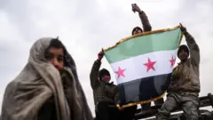 4 أشخاص يحمل ثلاثة منهم العلم السوري ذو النجوم الحمراء الثلاثة. يبدو أنهم يقفون في مشهد احتفالي. السماء ملبدة بالغيوم.