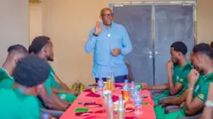 Umukuru w'ishyirahamwe ry'umupira w'amaguru mu Rwanda (FERWAFA), Fabrice Shema, ahagaze akoze ikimenyetso n'ikiganza, inyuma ye hari umuryango ufunze na serire. Imbere ye, ku mpande ebyiri z'ameza, hicaye abakinnyi b'Amavubi bambaye imyenda y'icyatsi kibisi, bamuteze amatwi, bamwe bamureba. Ku meza hari igitambaro gitukura. Ku meza hariho inyobwa byoroheje, by'amazi na Fanta. 