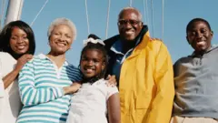 Uma família de quatro gerações posa para uma foto. Há uma menina, um homem na casa dos 20 anos, uma mulher de cerca de 40 anos e um casal de aproximadamente 60 anos. Eles estão em frente a um iate.