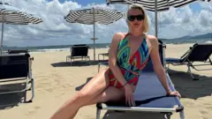 Anastasia Samsonova con lentes de sol y vestida con un colorido traje de baño está sentada sobre una silla para tomar sol en una playa. A su alredor las otras sillas con parasoles están vacías y se ve el mar de fondo