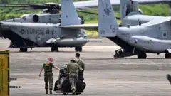 Três militares carregam equipamentos em pista com helicópteros de combate ao fundo