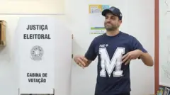 Fotografia colorida mostra Pablo Marçal dentro ao lado de cabine de votação usando uma camiseta e boné azuis com a letra M