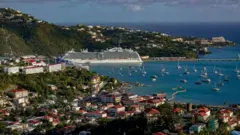 Panorama atual do porto de Saint Thomas, com pequenas embarcações e um grande navio de cruzeiro em um mar azul e um céu igualmente azul.