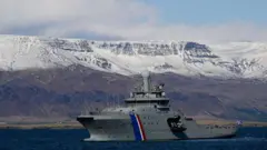 Uma embarcação da guarda costeira islandesa na água, com uma paisagem de montanhas nevadas ao fundo.