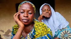 Aworan awọn iyawo ile meji, ọkan fọwọ lẹran, ọkan fọwọ sẹnu, wọn n woye .
Obinrin akọkọ wọṣọ alawọ ofeefe to ni omi aro, o we gele, o le tiroo soju, lo ẹgba ọrun, o lu imu rẹ pẹlu yẹti kekere, ṣugbọn o fọwọ lẹran bii ẹni to n ronu. Iya keji lo ibori alawọ omi aro, o wọ aṣọ alawọ kan naa, o fọwọ sẹnu, oun naa n wo orere aye.