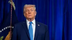 Imagen de donald trump. Viste un traje azul y corbata del mismo color. Está de pie, con rostro serio.
