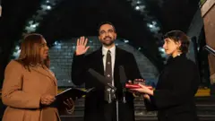 Zoran Mamdani, de barba, corbata y abrigo oscuro, levanta la mano derecha al ser juramentado por la fiscal general de Nueva York Letitia James (a la izquierda) que sostiene un documento. Rama Duwaji (a la derecha), una mujer con abrigo oscuro lo mira atentamente y sostiene un Corán donde Mamdami apoya su mano izquierda.