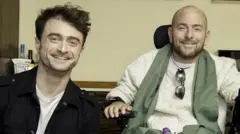 Daniel Radcliffe w białym topie i czarnej kurtce obok Davida, który ma na sobie kremowy sweter i porusza się na wózku inwalidzkim. Obaj uśmiechają się, siedząc w środku.