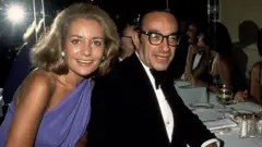 Barbara Walters y Alan Greenspan son fotografiados en una cena de etiqueta en 1977. Miran a la cámara y sonríen. Walters lleva un vestido azul y Greenspan viste un esmoquin.