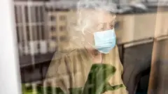 Mulher idosa de máscara olhando pelo vidro de um janela