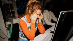 A woman in an orange long-sleeve shirt and an argyle vest sits at her work desk with her head in her hands. 