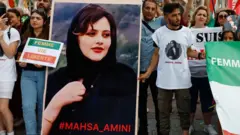 Uma pessoa carrega um cartaz no qual está escrito "Mahsa_Amini" em vermelho, com a imagem de Amini. O cartaz é cercado por homens e mulheres usando camisetas com a imagem dela e carregando bandeiras para marcar o primeiro aniversário de sua morte.
