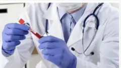 Ọwọ to mu ike ẹjẹ dani, to n yẹ ẹjẹ inu rẹ wo. Ẹni naa wọ aṣọ funfun, o fi ibomu bo imu rẹ o si lo ibọwọ si ọwọ rẹ mejeeji. Bakan naa lo fi irinṣẹ ayẹwo awọn dokita kọ ọrun rẹ.