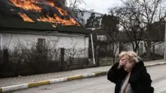 زنی مقابل خانه‌ای در حال سوختن در ایرپین، خارج از کی‌یف ایستاده و صورت خود را با دست پوشانده است، ۴ مارس ۲۰۲۲