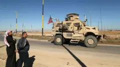 Üzerinde ABD bayrağı olan bir zırhlı araç otoyolda ilerliyor. İki adam aracın geçişini izliyor.