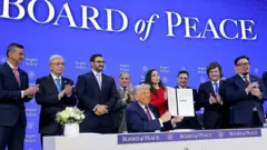 الرئيس الأمريكي دونالد ترامب وهو جالس يحمل وثيقة عليها توقيعه حول "مجلس السلام" في غزة وأمامه شريط كُتب عليه عبارة: "Board of Peace- مجلس السلام".
