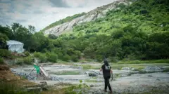 Uma pedreira com vegetação ao fundo. Uma auditora fiscal do trabalho caminha pelo cenário